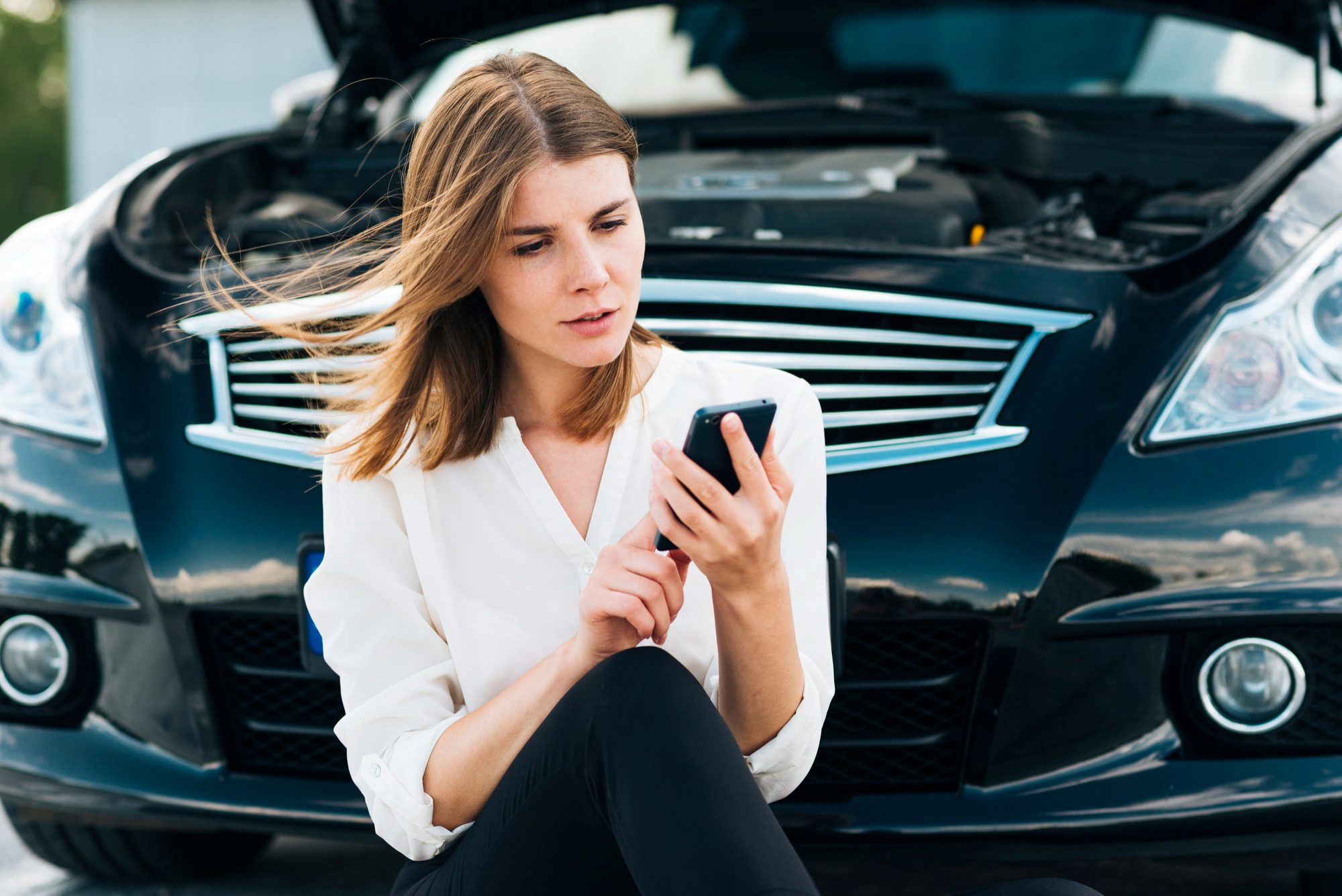Sachmängelhaftung bei Gebrauchtwagen, Besorgte Frau sitzt vor einem schwarzen Auto mit geöffneter Motorhaube und nutzt ihr Smartphone