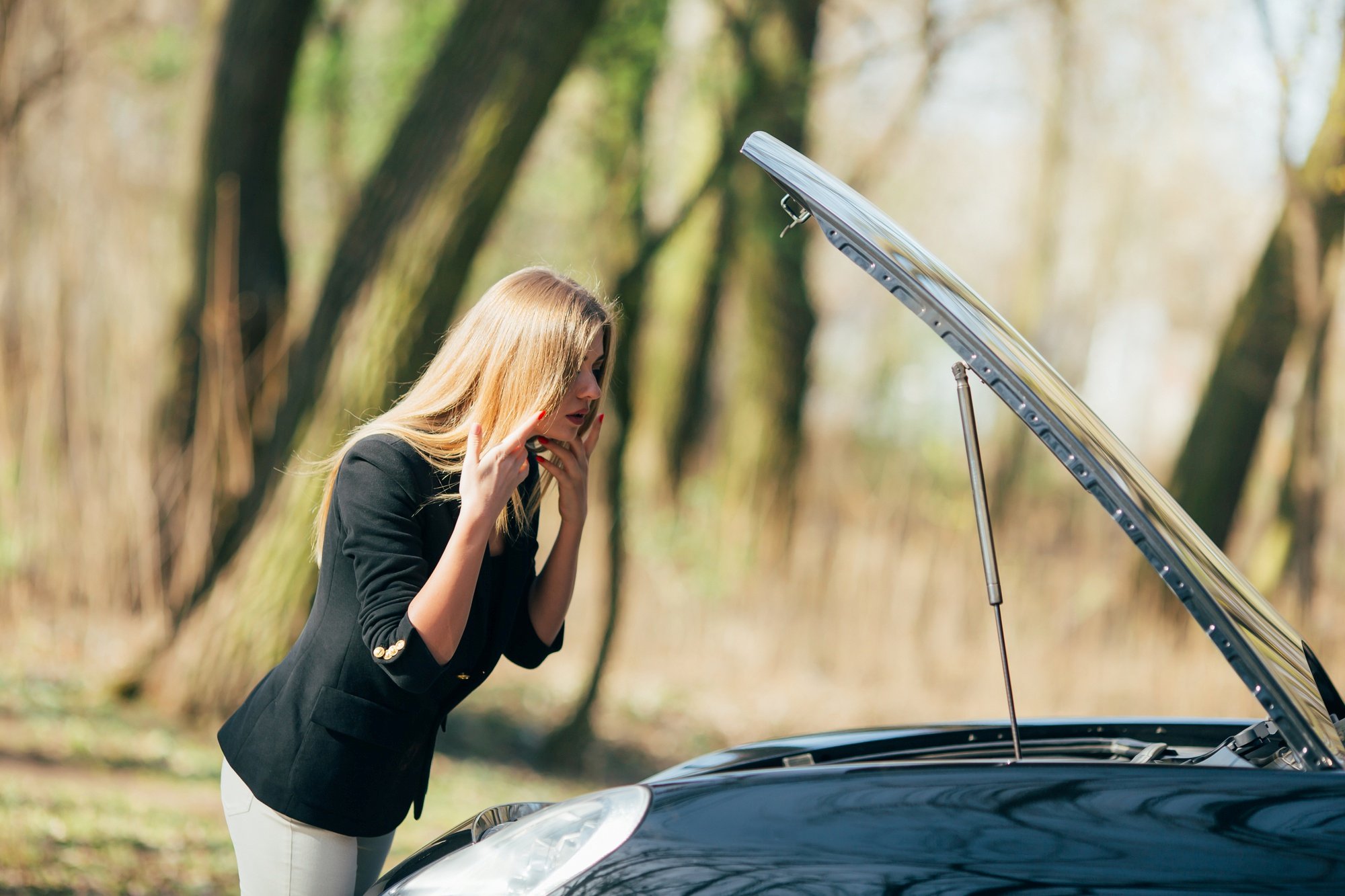 Sachmängelhaftung beim Gebrauchtwagen Besorgte Frau steht vor einem schwarzen Auto mit geöffneter Motorhaube und nutzt ihr Smartphone.